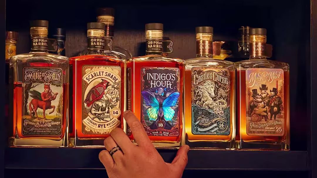 A hand reaches for one of five bottles of whiskey with ornate labels, displayed side by side on a shelf.