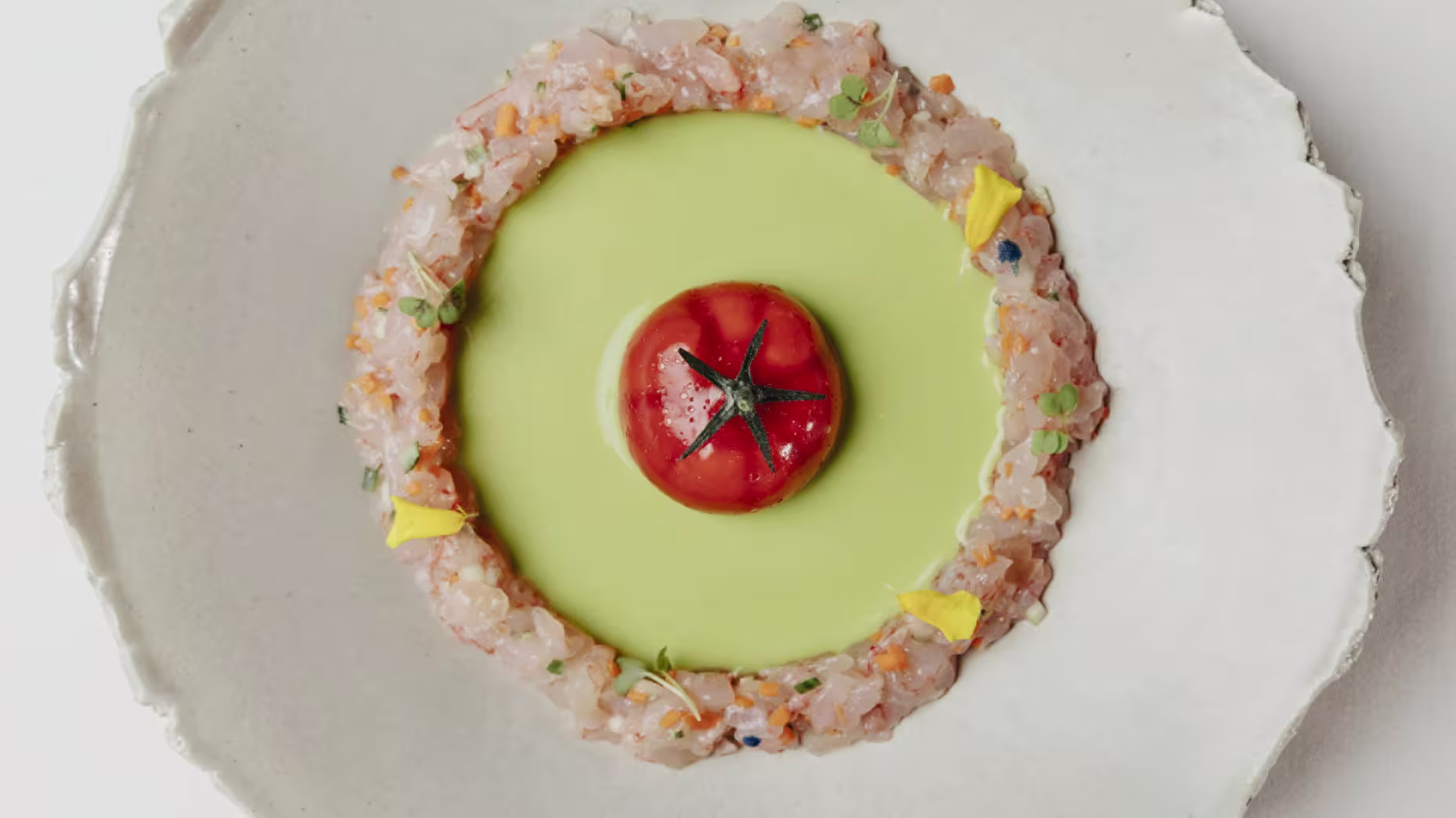 Dani Brasserie presenta una obra maestra culinaria con un tomate glaseado al centro, rodeado por crema verde y tartar de mariscos finamente picado.