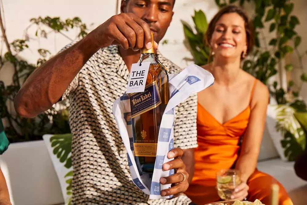 A man unwraps a bottle of Johnnie Walker Blue Label whisky while a woman in an orange dress smiles and holds a drink, both seated outdoors.