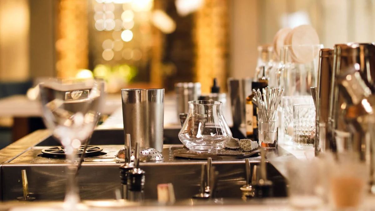 A close-up of a bar counter with cocktail shakers, glassware, metal tools, and bottles, set in a softly lit environment.