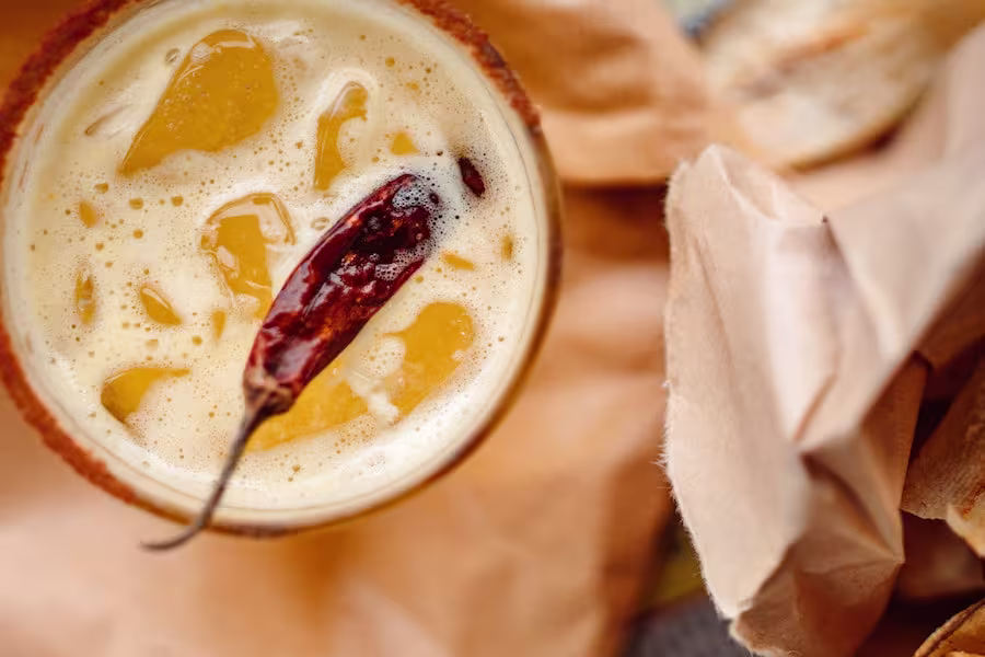 A frothy drink topped with a dried chili pepper, viewed from above, next to a piece of crumpled brown paper.