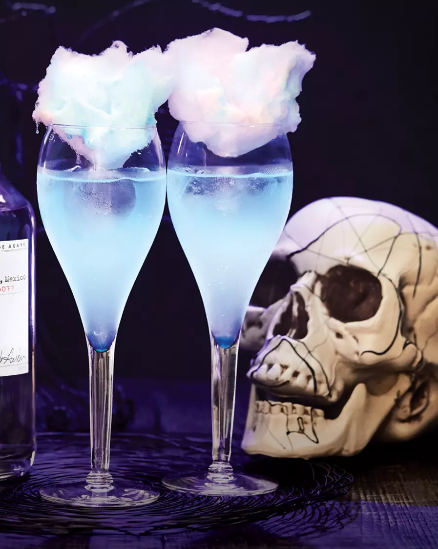 Two blue cocktails in wine glasses, each topped with cotton candy, are placed beside a white decorative skull and a bottle, against a dark background.