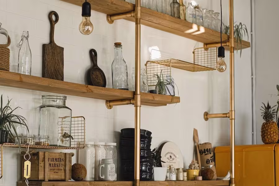 Open wooden shelves with glass jars, mugs, plates, plants, cutting boards, and hanging light bulbs against a white tiled kitchen wall.