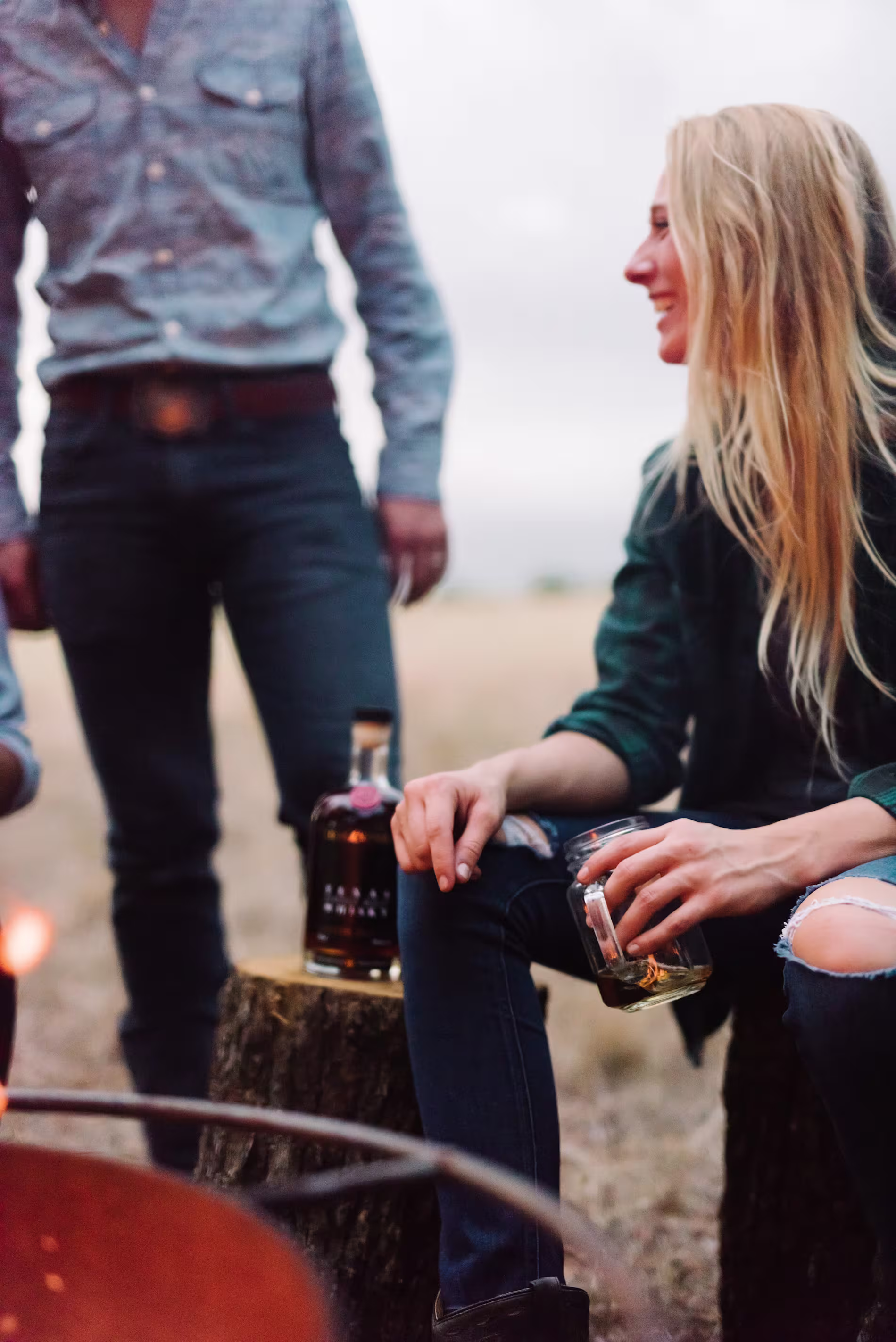 Two people sit outdoors by a fire; one woman holds a glass and smiles while a bottle sits on the tree stump between them.