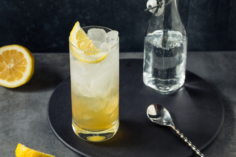 A glass of iced drink with a lemon wedge, a spoon, a bottle of clear liquid, and lemon slices on a dark round tray.