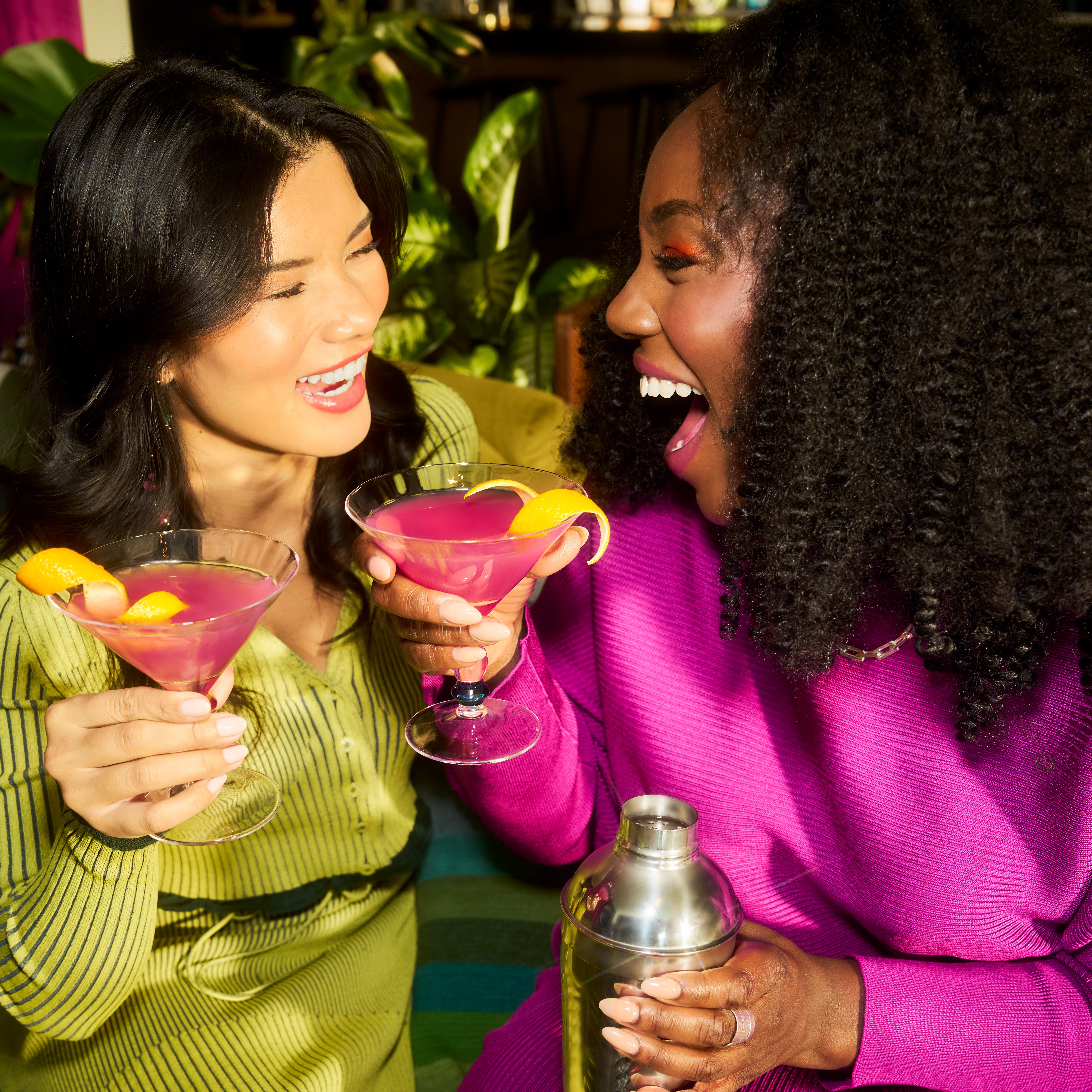 Două femei râd una de cealaltă în timp ce țin cocktailuri în mână.