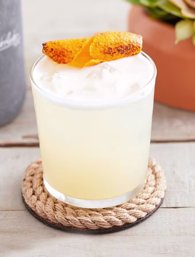 A tequila cocktail with burnt orange sits on a woven drinks mat on a wooden table.