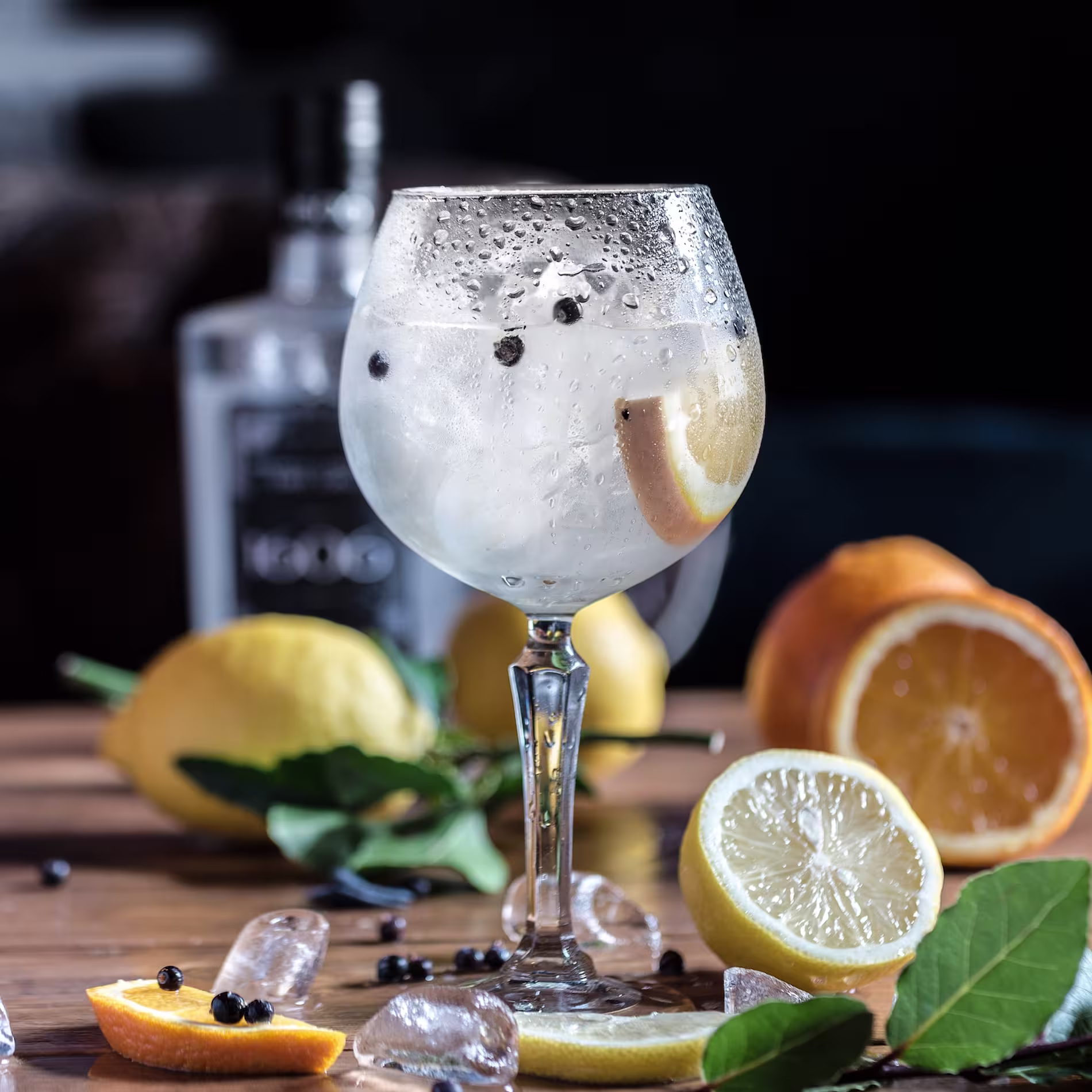 A gin and tonic with ice, lemon slice, and juniper berries in a stemmed glass, surrounded by lemons, oranges, ice cubes, and a bottle on a wooden table.