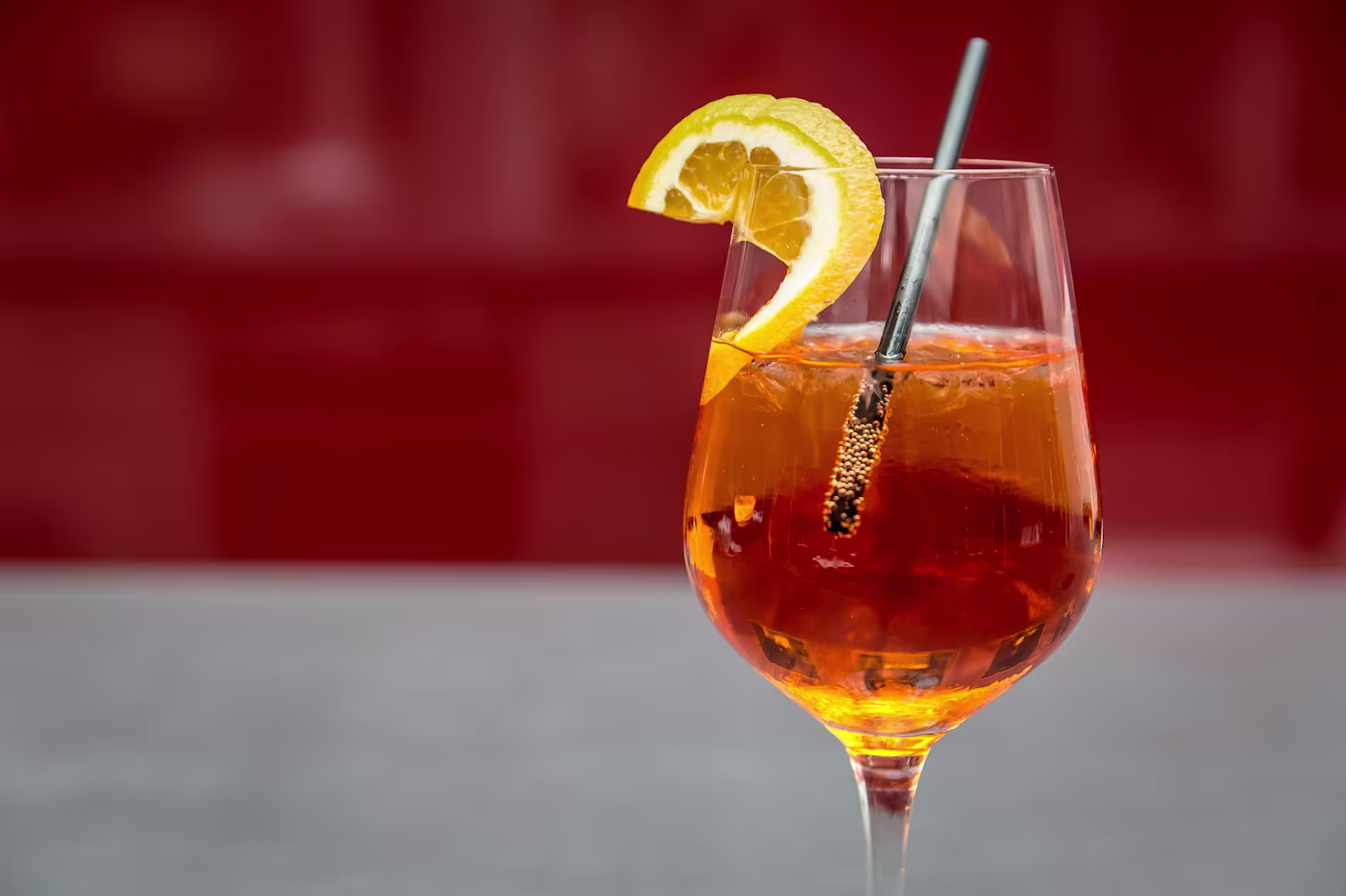 A wine glass filled with an orange-colored drink, ice cubes, a metal straw, and garnished with a slice of orange, set against a blurred red background.
