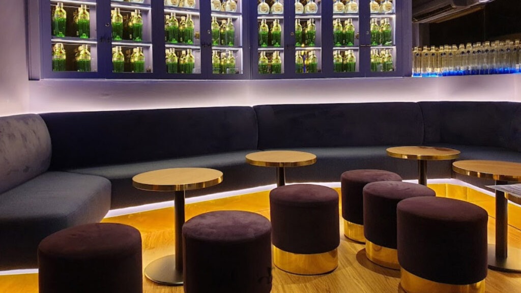 Zona de sofás en Gin Fizz Bilbao Cocktail Bar con botellas iluminadas en vitrinas, mesas redondas doradas y taburetes tapizados a juego.