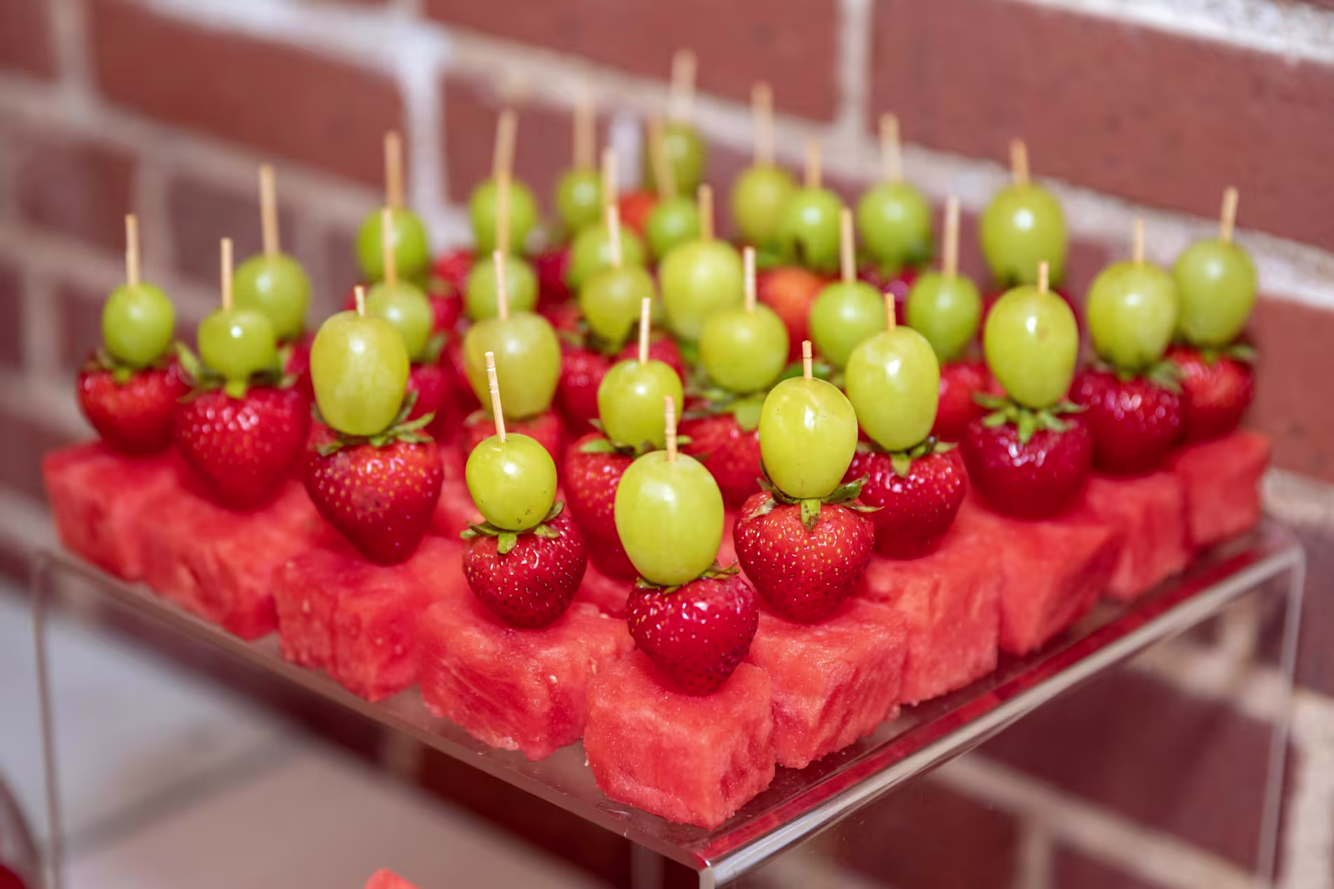 Skewers with green grapes and strawberries are arranged on cubes of watermelon, displayed on a clear stand against a brick wall background.