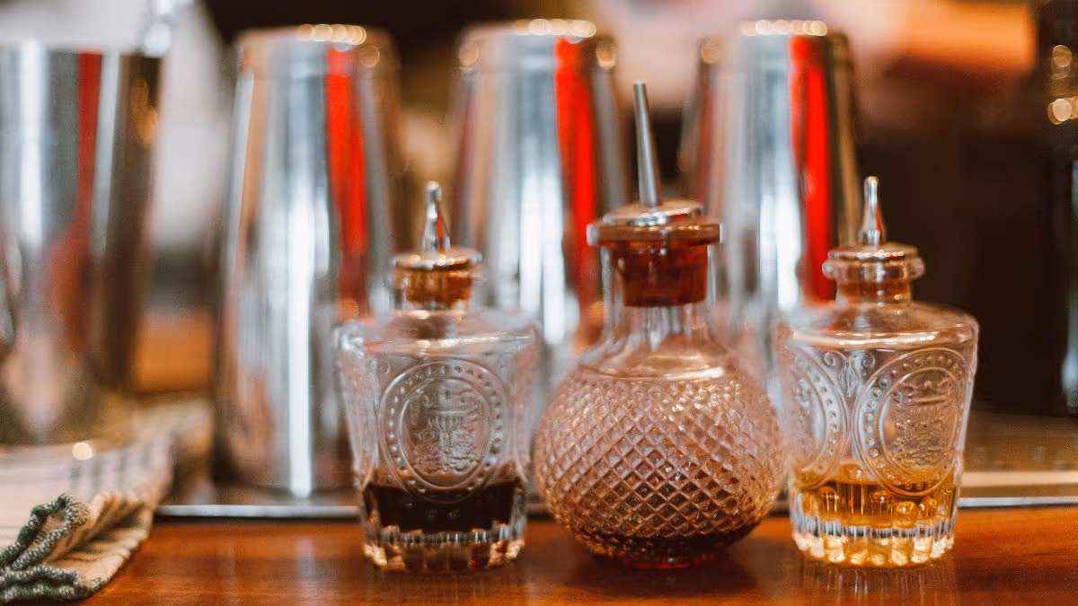 Three glass bottles with stoppers, containing different colored liquids, are placed on a bar counter in front of metal cocktail shakers.