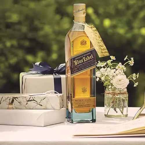 A bottle of Johnnie Walker Blue Label whisky stands on a table next to wrapped gifts, a small flower bouquet in a jar, and an open notebook, with greenery in the background.