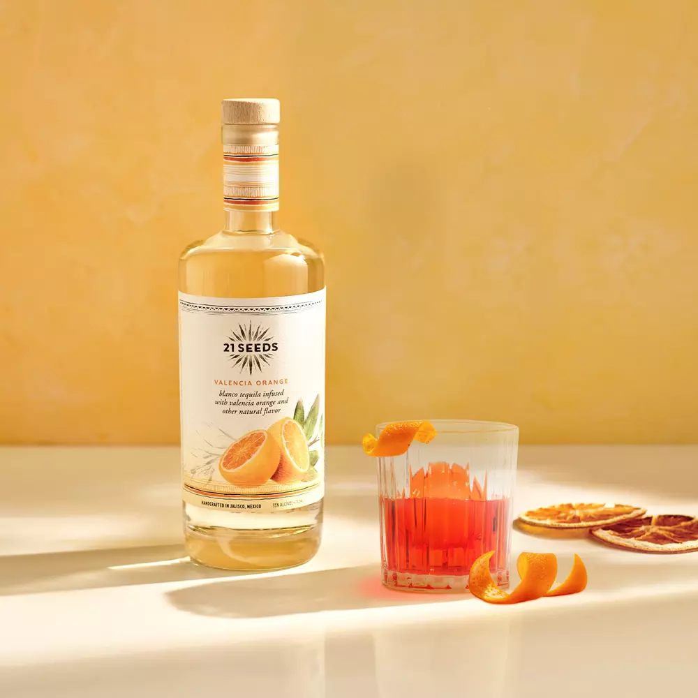 A bottle of 21 Seeds Valencia Orange tequila stands next to a glass of orange cocktail with orange garnishes and dried orange slices on a light surface against a yellow background.