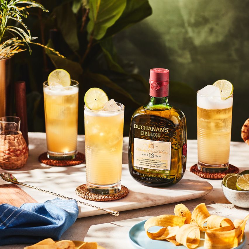 La imagen muestra una botella de whisky Buchanan's de 12 años junto a tres vasos con cócteles preparados con dicho destilado, rodeados de elementos de coctelería y alimentos.