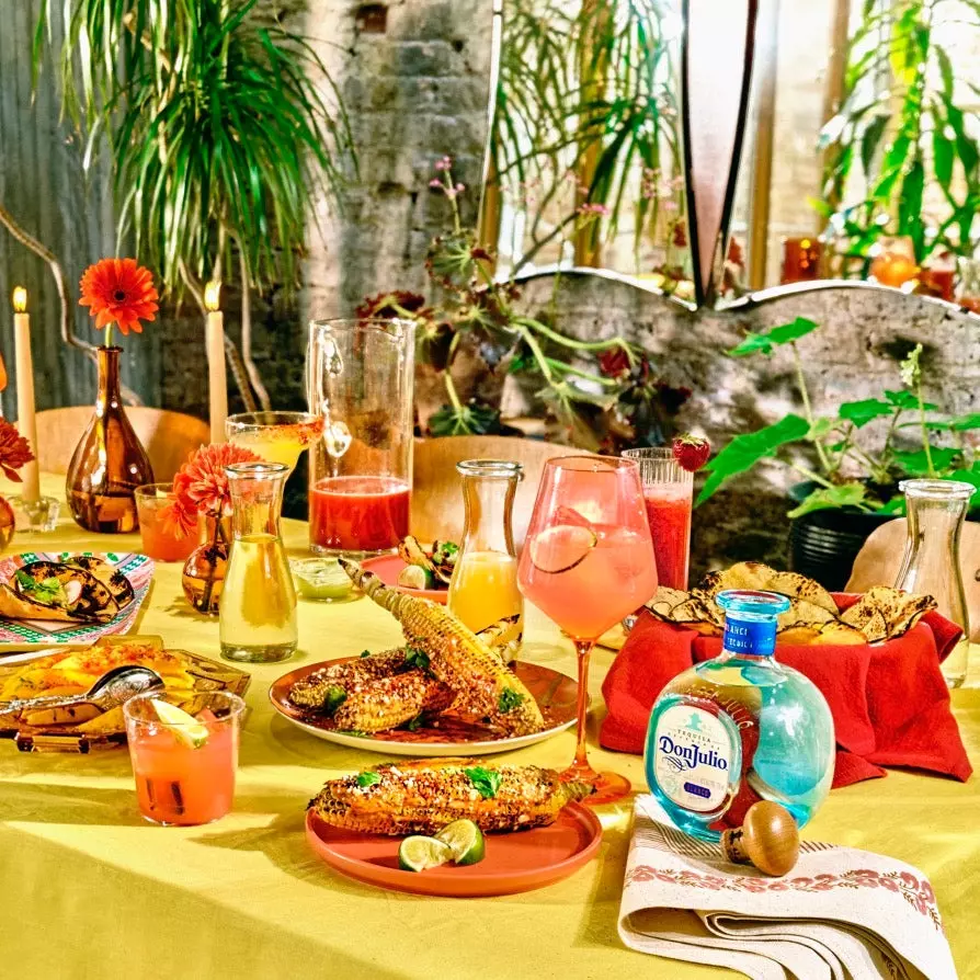 A festive table set with Mexican food, colorful drinks, grilled corn, candles, a bottle of Don Julio tequila, and vibrant decorations, surrounded by plants.