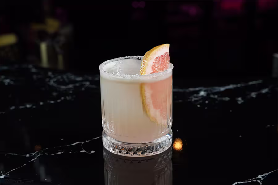 A glass with a salted rim contains a pale cocktail garnished with a wedge of grapefruit, set on a dark marble countertop.