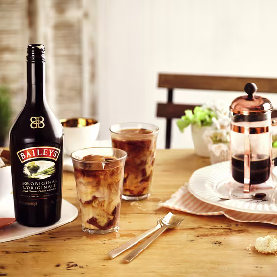 La imagen muestra una botella de licor de crema Baileys junto a dos vasos con cócteles preparados con este licor, una cafetera y otros elementos de coctelería.