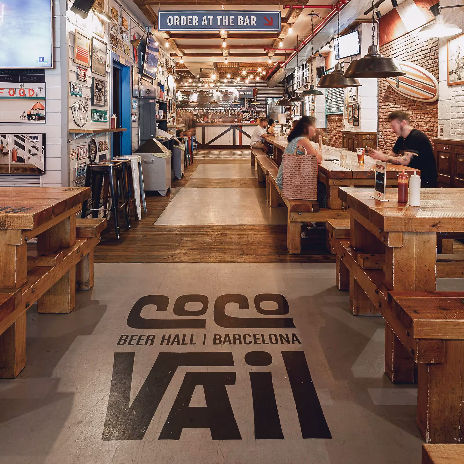 La imagen muestra el interior del Cocovail con una estética industrial. Hay un largo pasillo que conduce a la zona de la barra, que tiene un letrero que dice “ORDER AT THE BAR”. En el suelo, hay texto pintado que dice “COCO BEER HALL | BARCELONA”