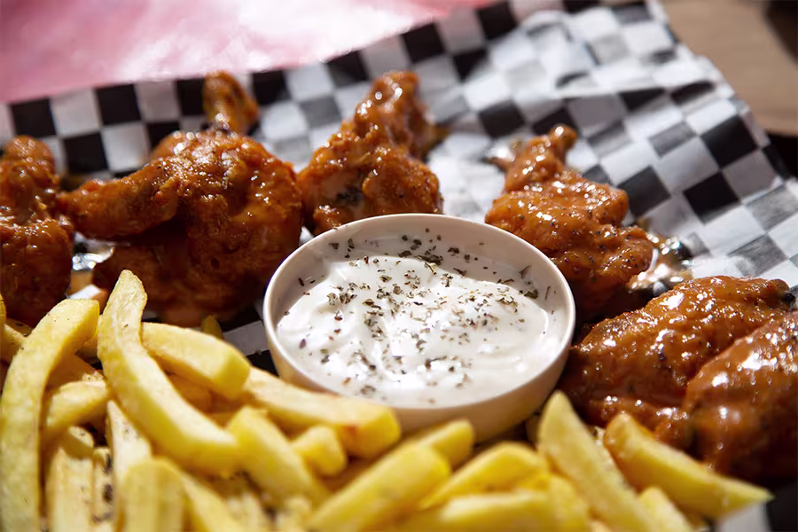 Chicken wings coated in sauce and French fries are served on checkered paper with a bowl of ranch dipping sauce topped with herbs.