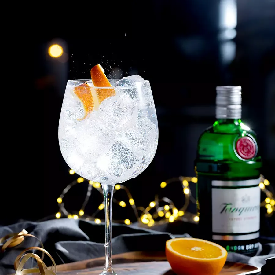 A glass of gin and tonic with ice and an orange twist sits on a wooden board beside a green Tanqueray gin bottle and a halved orange, with string lights in the background.