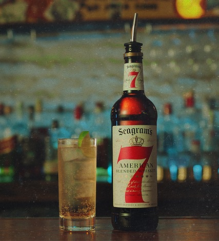 A bottle of Seagram’s 7 Crown whiskey and a mixed drink with ice and lime sit on a bar counter with blurred liquor bottles in the background.
