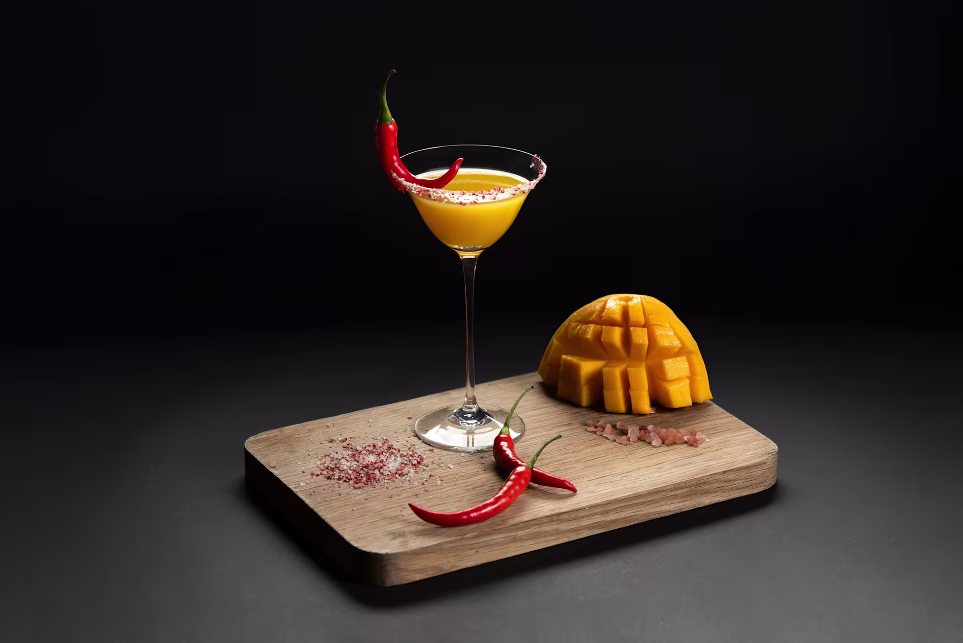 A martini glass with a yellow cocktail garnished with a red chili, next to a sliced mango, red chilies, and coarse salt on a wooden board against a dark background.