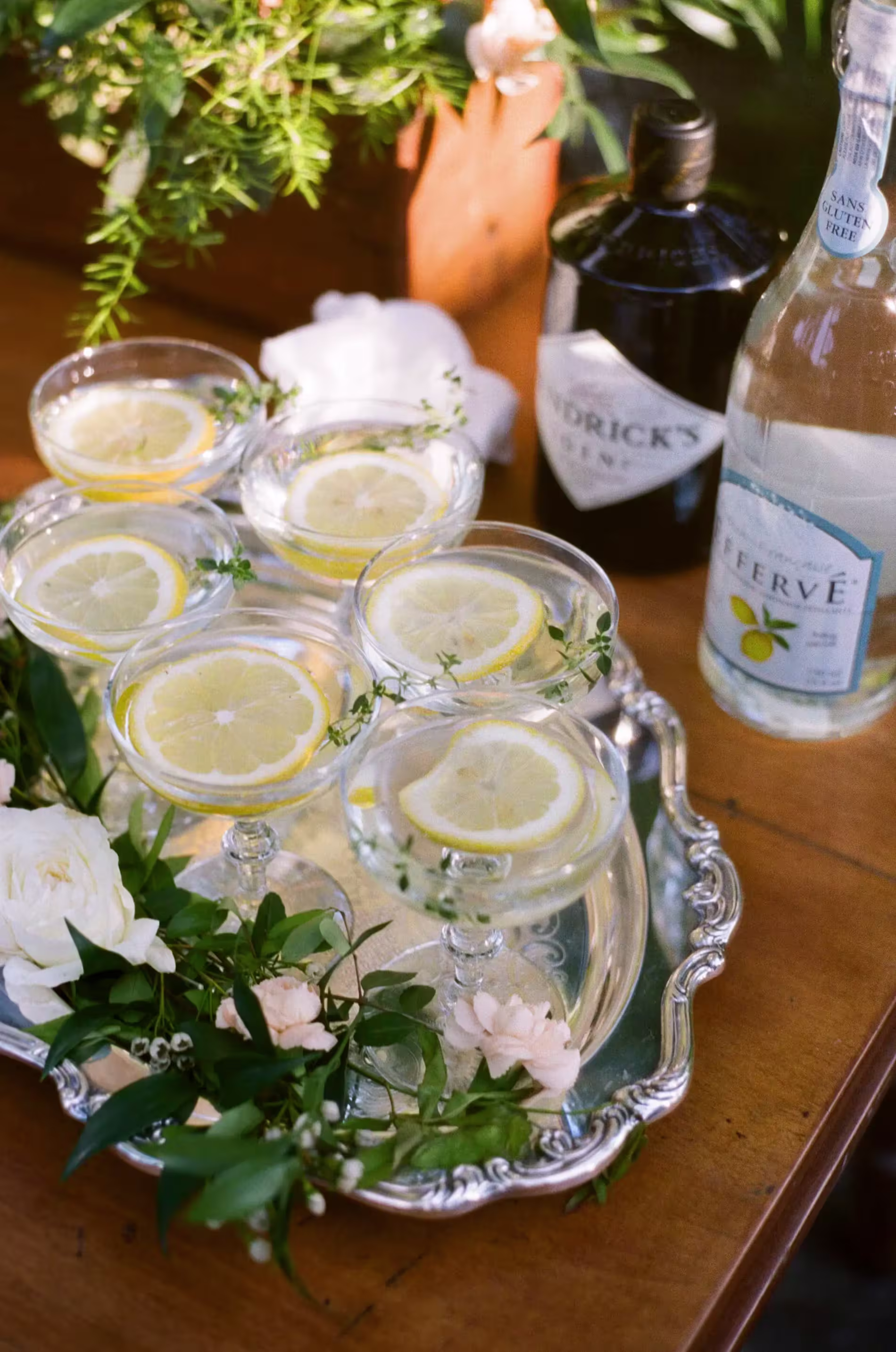 A silver tray holds six cocktails garnished with lemon slices and herbs, surrounded by flowers, with gin and sparkling water bottles nearby.