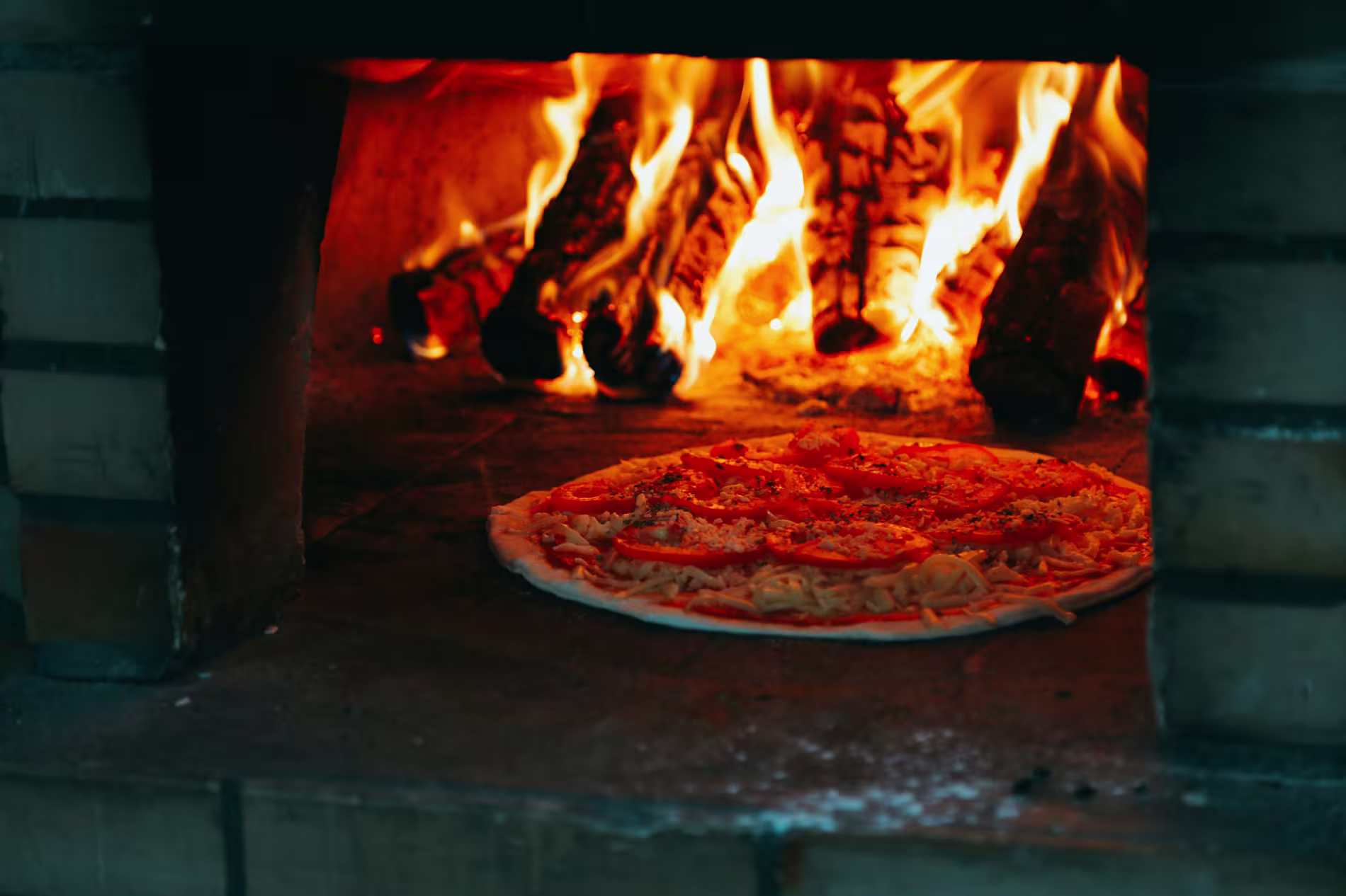 Uncooked pizza with toppings on a stone surface baking in a wood-fired brick oven with burning logs and visible flames.