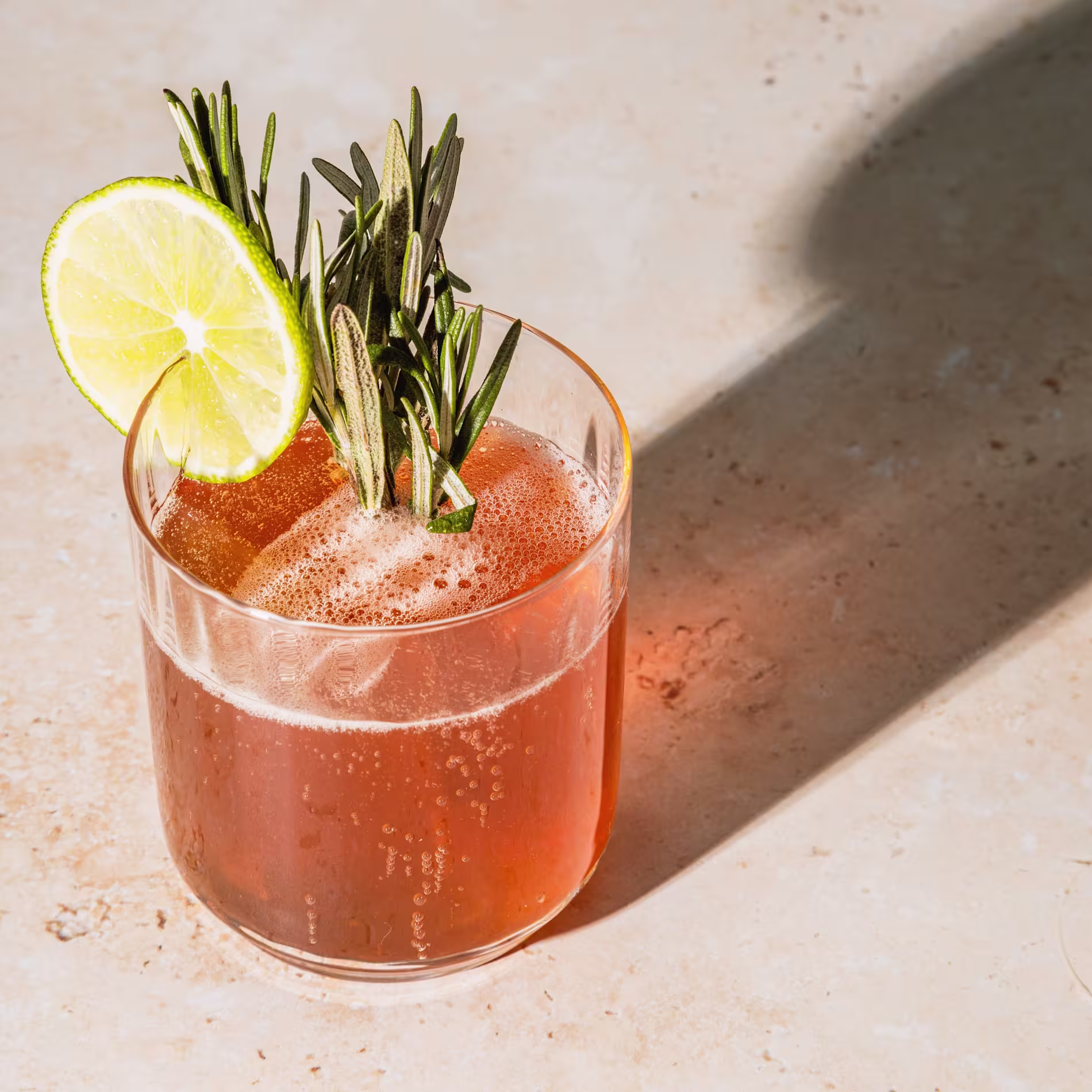 A glass of reddish-brown bubbly drink garnished with a lime slice and sprigs of rosemary on a light-colored surface.