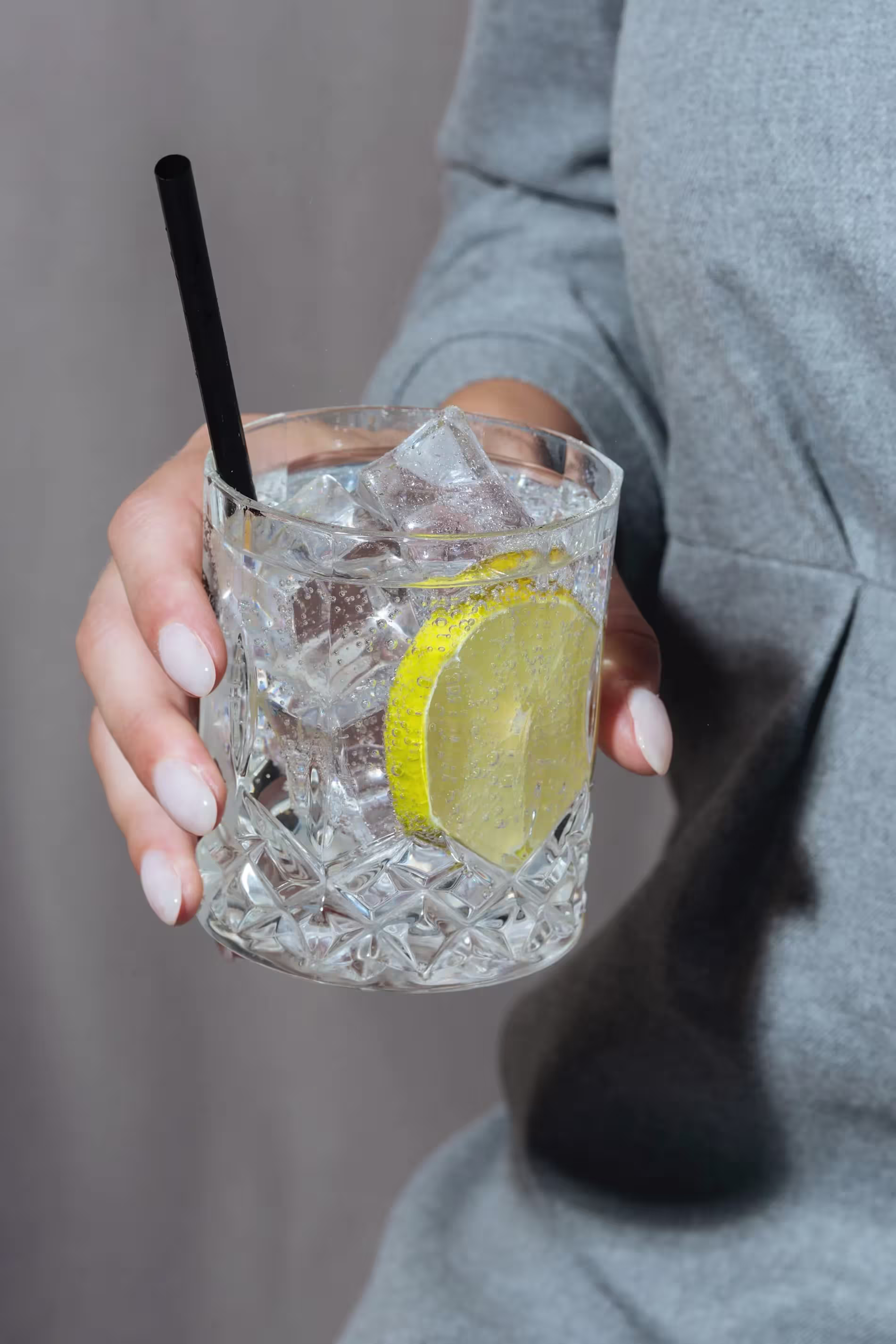 A person holding a glass of iced water with a lemon slice and a black straw, wearing a grey long-sleeve top.