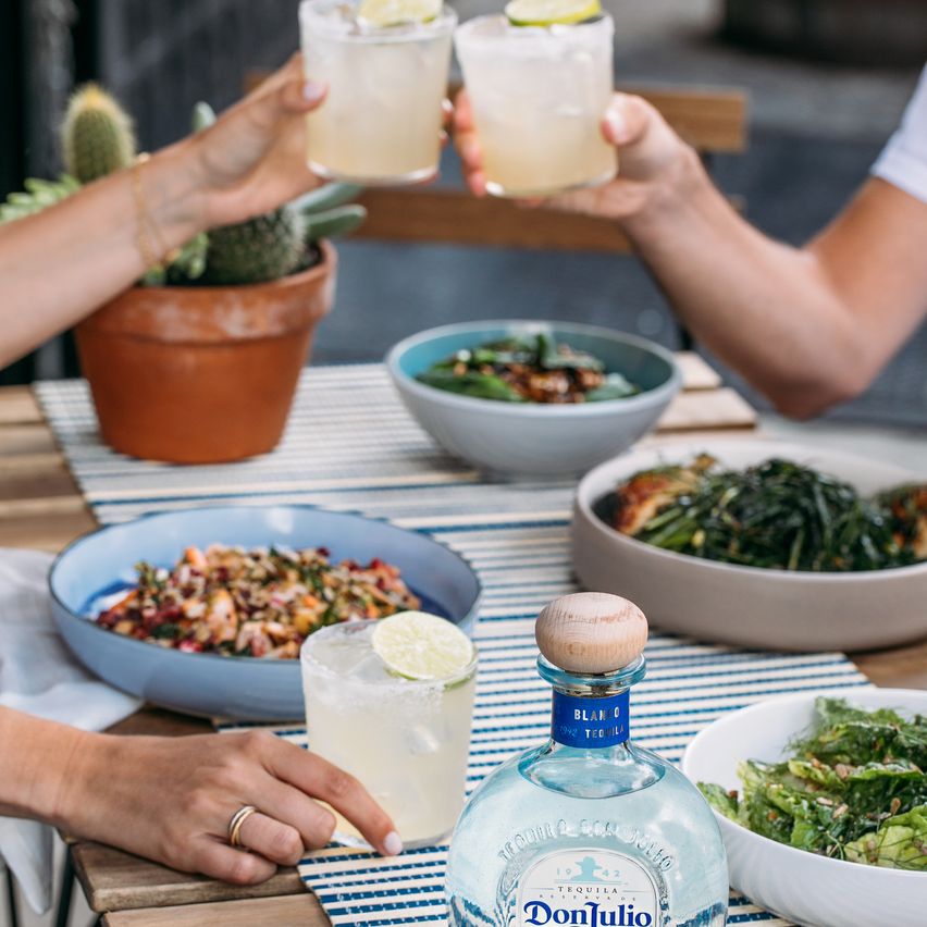 La imagen muestra una mesa con una variedad de platos de comida, destacando en el centro una botella de tequila Don Julio Blanco, mientras que dos personas brindan con cócteles Margarita.