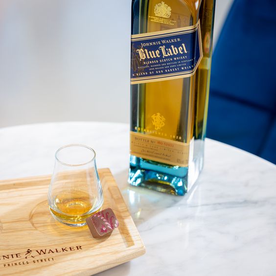 La imagen muestra una botella de whisky Johnnie Walker Blue Label junto a un vaso que contiene este destilado, con un chocolate ubicado a un lado.