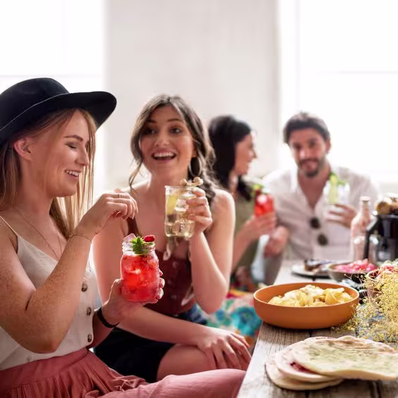 La imagen muestra cuatro personas, cada una con un cóctel en la mano, junto a una mesa con diversos platos de comida.