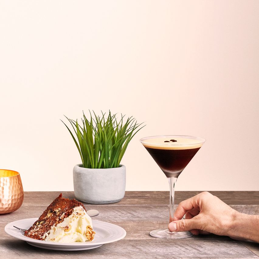 En la imagen se puede observar una copa que contiene el cóctel Espresso martini, acompañada por un plato con una porción de torta, y una maceta con una planta verde como decoración.