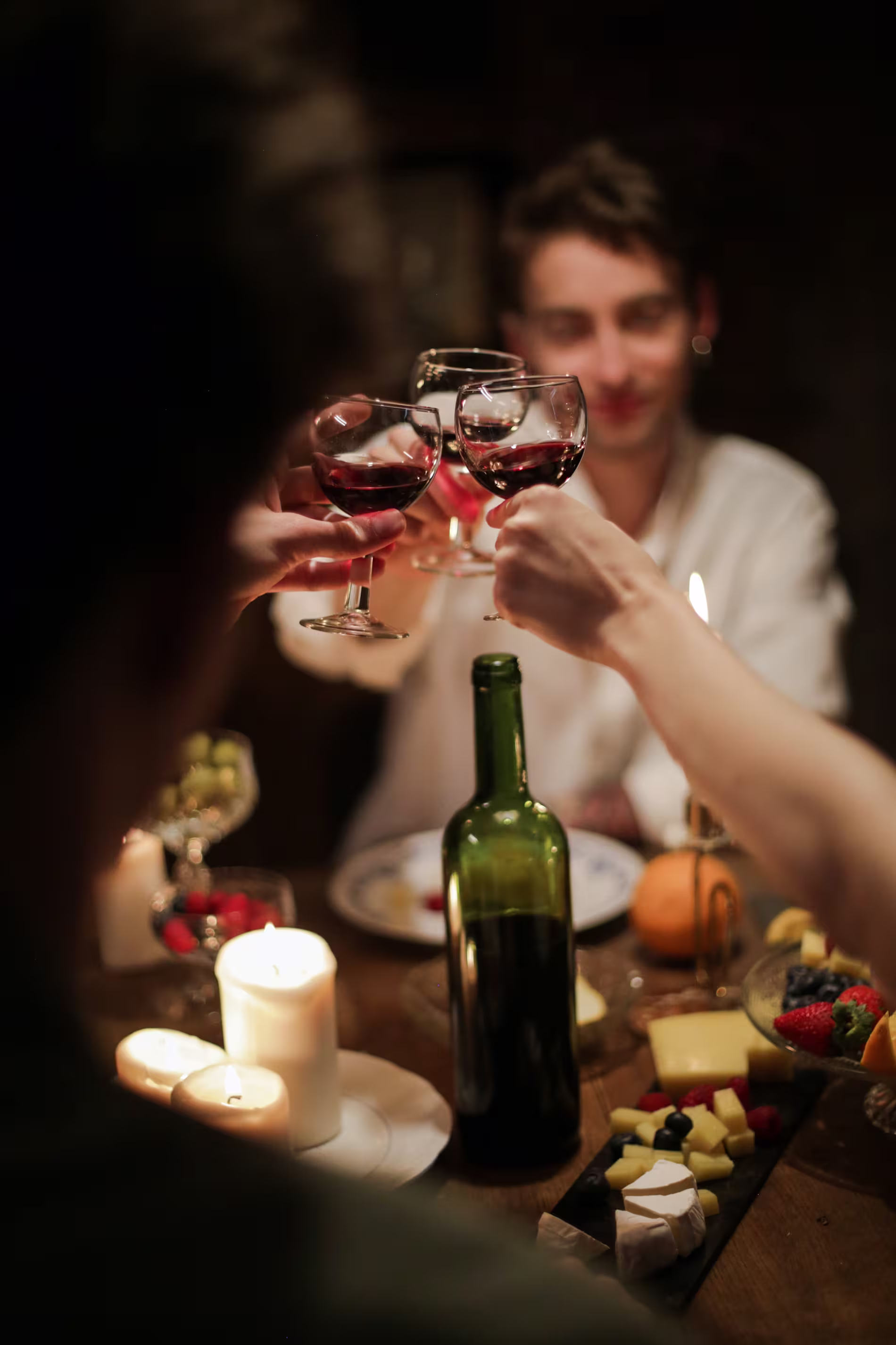 Three people clink glasses of red wine at a candle-lit dinner table with a bottle of wine, cheese, fruit, and candles.