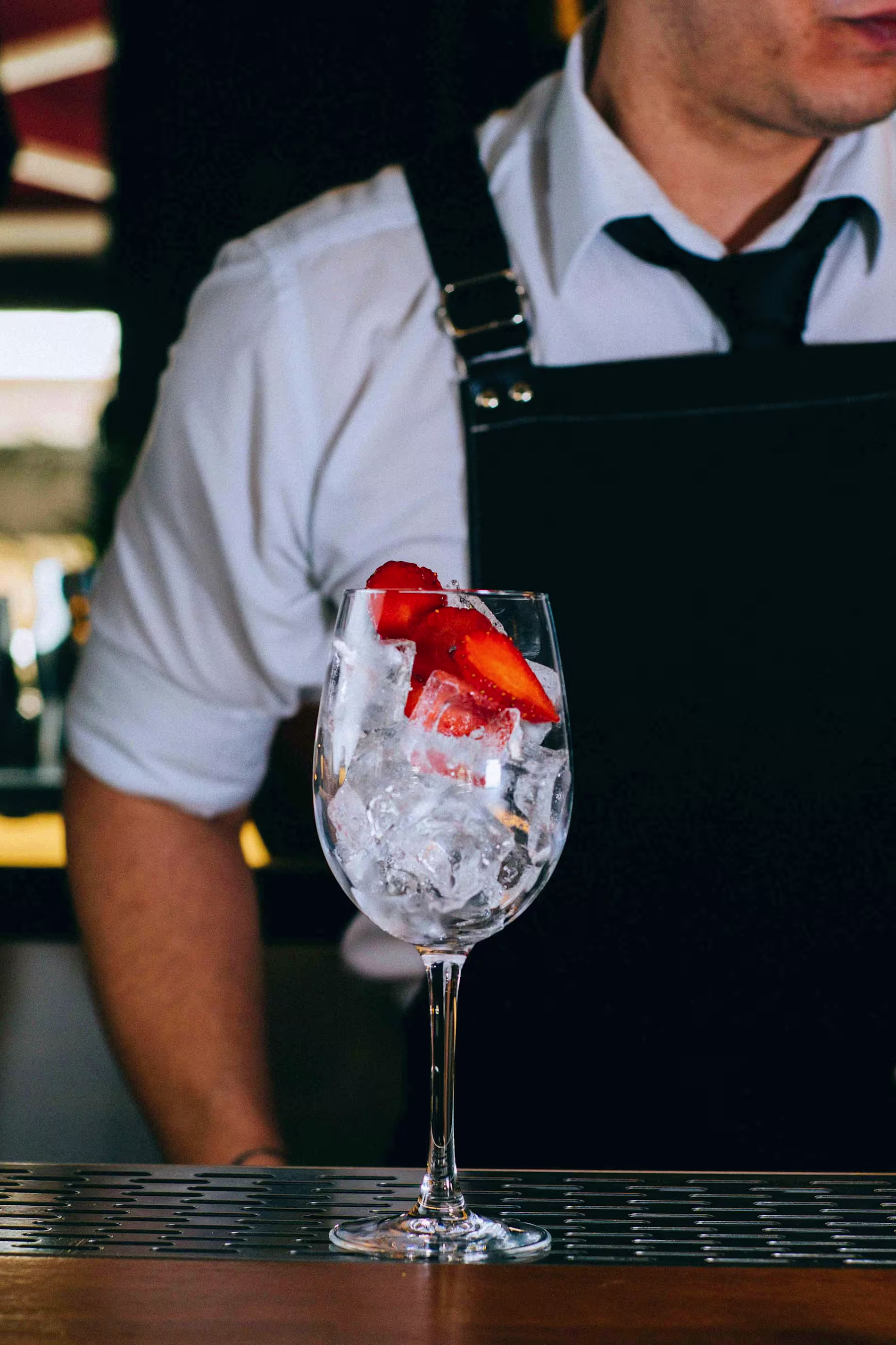 A bartender in a white shirt and black apron stands behind a wine glass filled with ice and sliced strawberries on a bar counter.