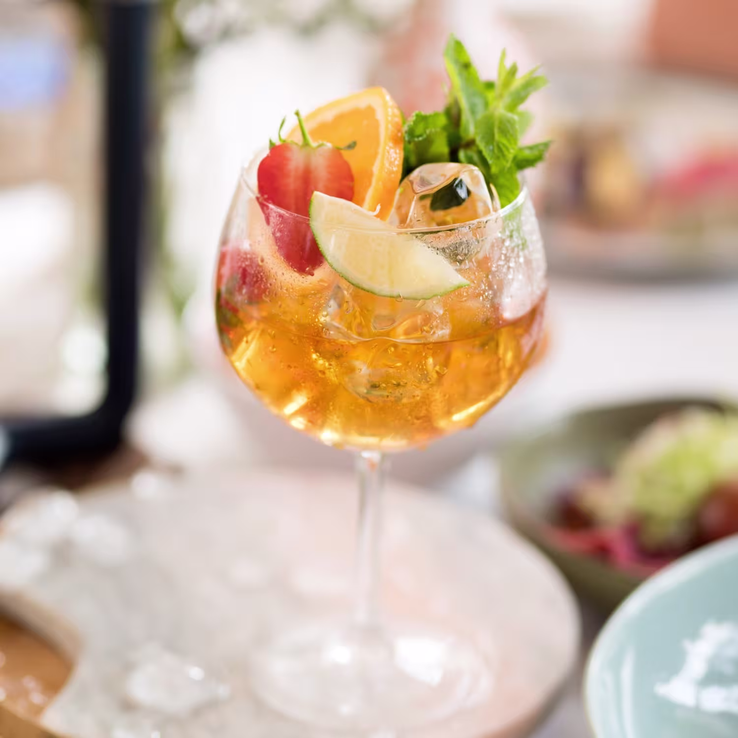 Un cóctel en un vaso grande con hielo, adornado con una fresa, una rodaja de naranja, un gajo de lima y una ramita de menta, sobre una superficie de mármol.