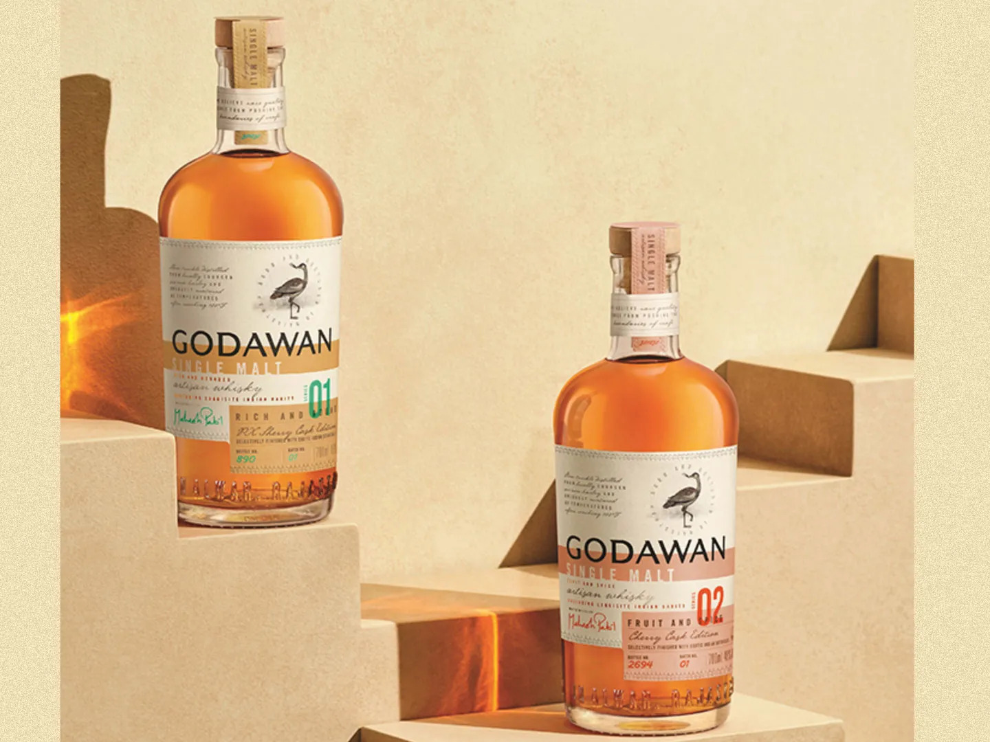 Two bottles of Godawan single malt whisky, labeled 01 and 02, are displayed on beige geometric steps against a neutral background.