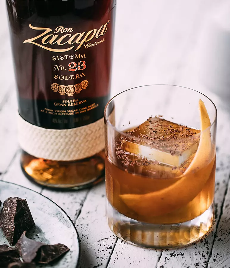 A glass of rum cocktail with a large ice cube, orange peel, and chocolate shavings sits next to a bottle of Ron Zacapa 23 on a white wooden surface.