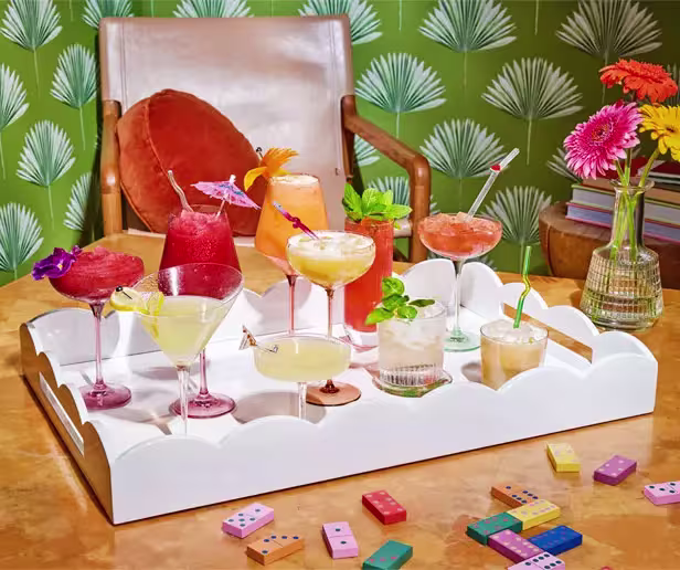 A white tray holds various colorful cocktails in glasses, garnished with fruit and umbrellas, on a table with dominoes; a chair and vase of flowers are in the background.
