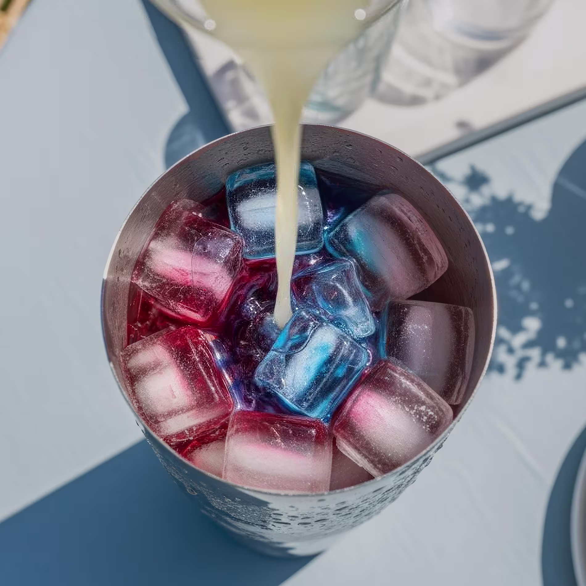 Élénk színű folyadékokat öntenek felülről egy jéggel töltött ezüst koktélkeverőbe.