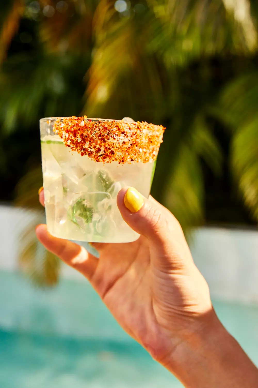 A hand with yellow nail polish holds a glass of iced beverage with a chili powder rim, outdoors near a pool and palm trees.