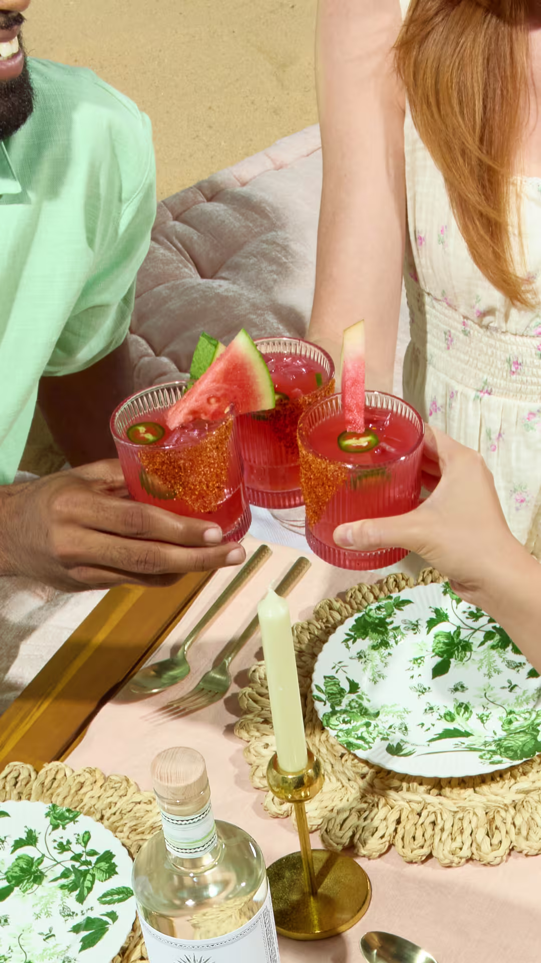 Three people toast with pink cocktails garnished with watermelon and jalapeño at an outdoor picnic with floral plates and a lit candle on a sandy beach.