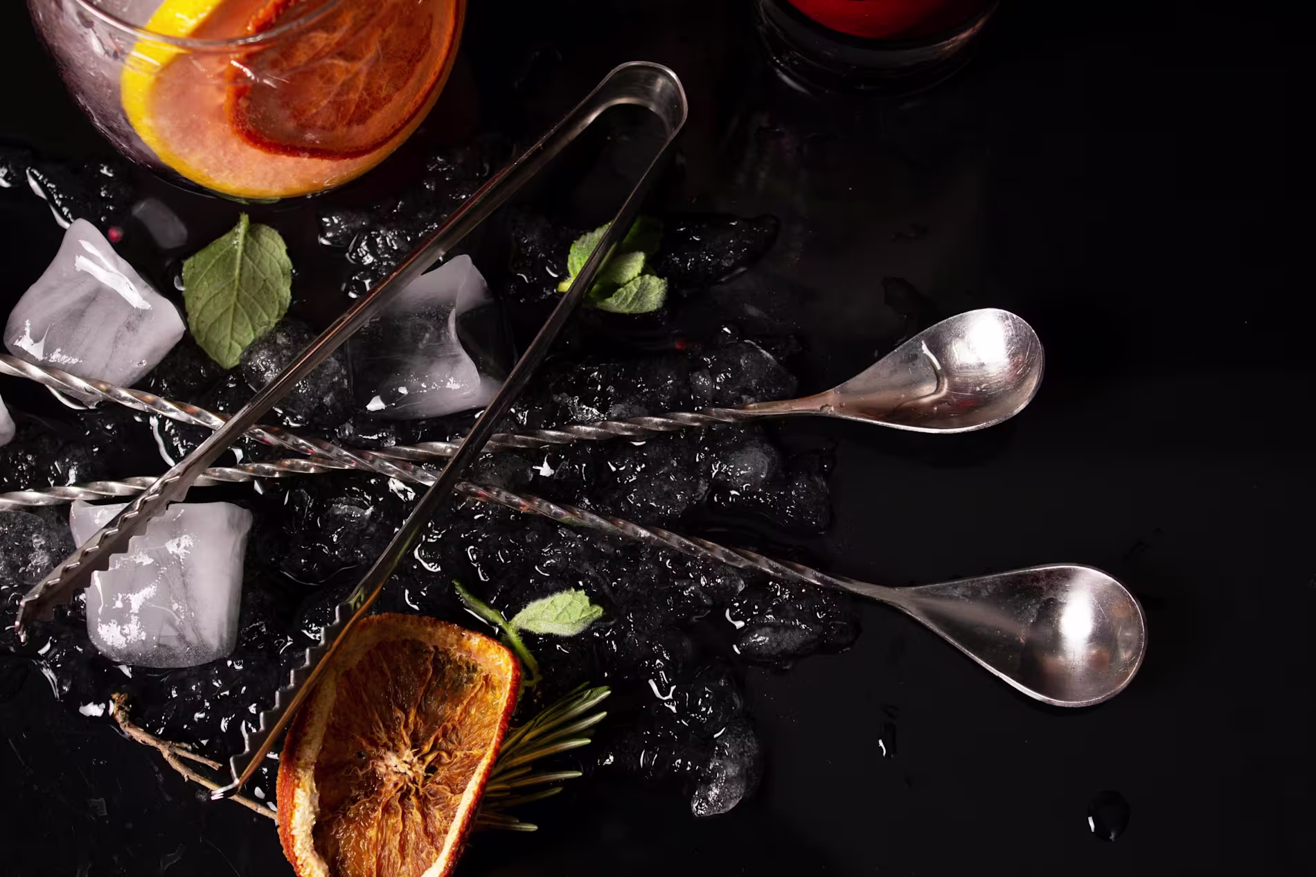 Two metal bar spoons, ice cubes, mint leaves, and dried orange slices are arranged on a black surface with a cocktail partially visible in the corner.