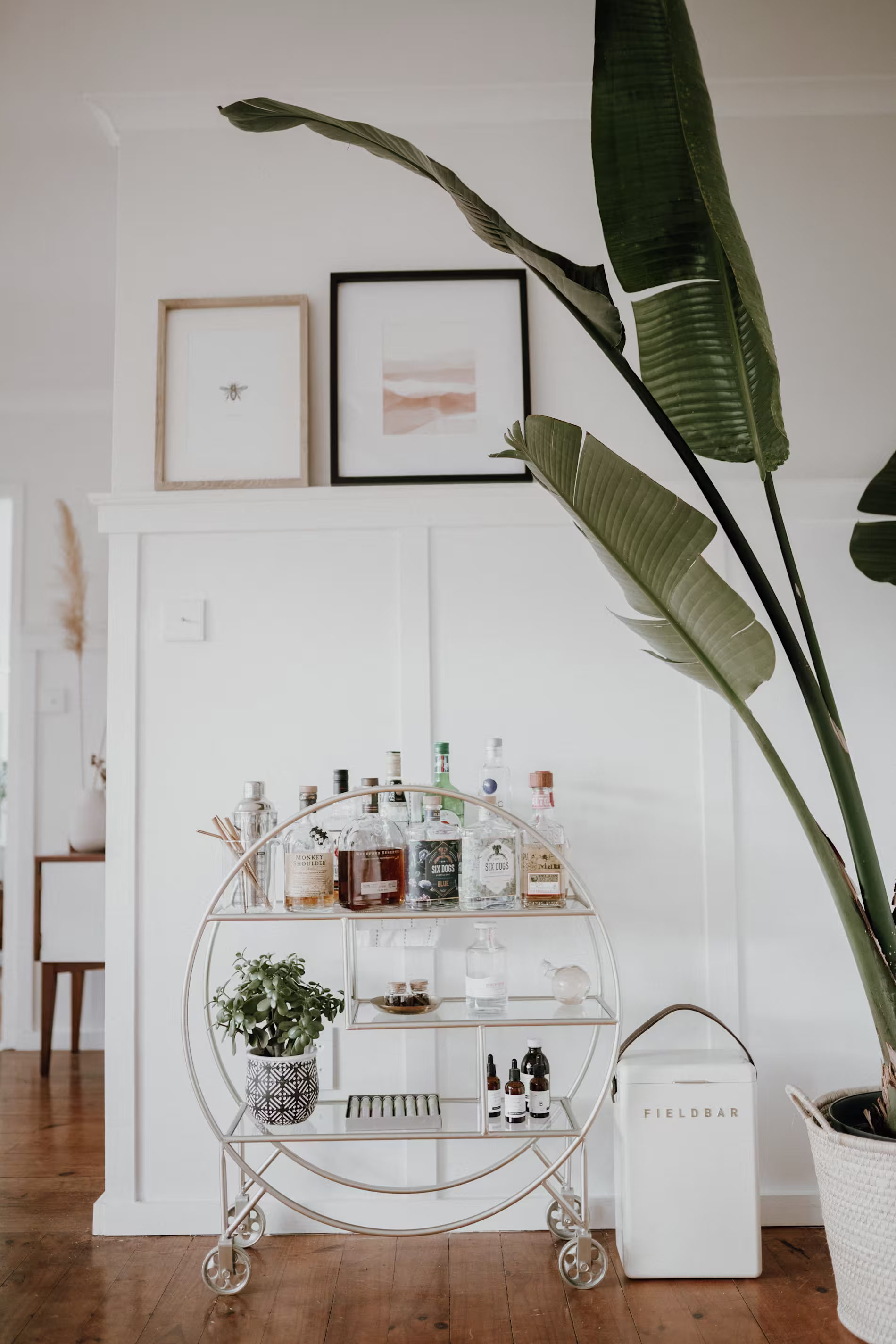 A circular bar cart with assorted bottles, plants, and glassware stands against a white wall with framed art, beside a tall potted plant on a wooden floor.