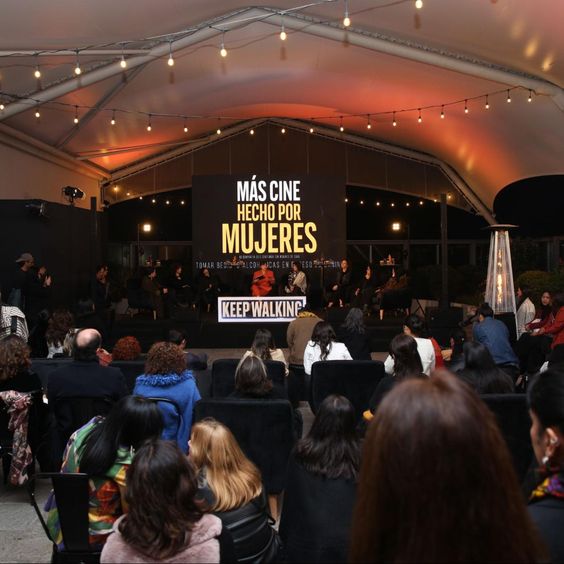 La imagen muestra un foto panorámica del conversatorio y, de fondo, una pantalla con el texto: Más cine hecho por mujeres.