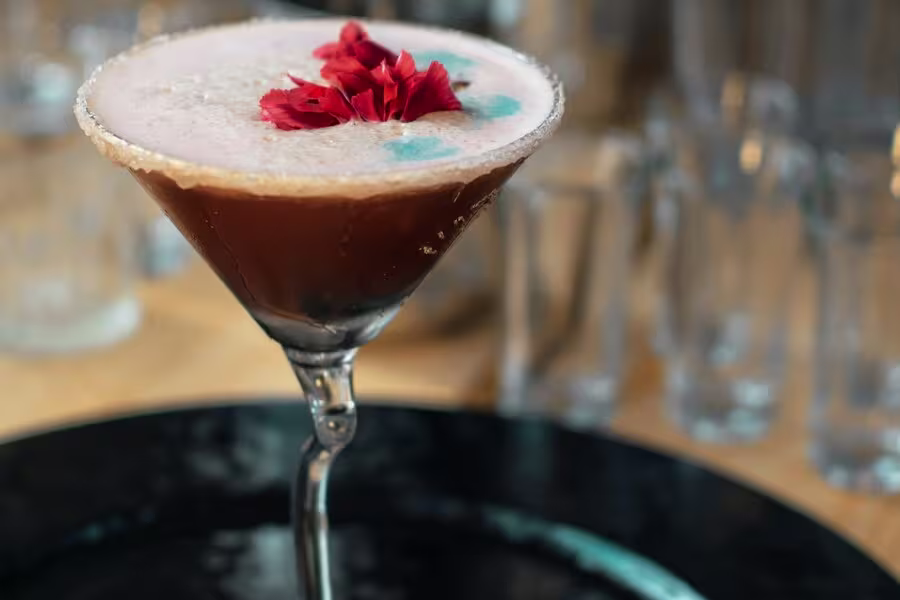 A brown cocktail in a martini glass with a sugared rim, frothy top, blue spots, and red flower petals as garnish, set on a black plate with glassware in the background.