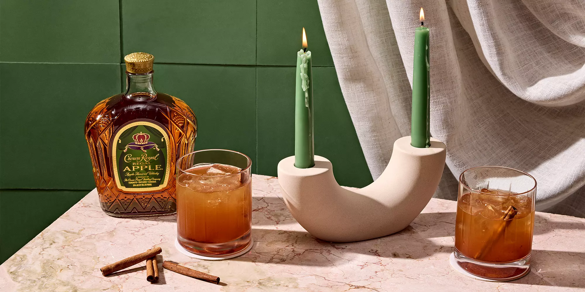A bottle of Crown Royal Apple, two cocktails with ice, a beige candle holder with two green candles, and cinnamon sticks on a marble surface.