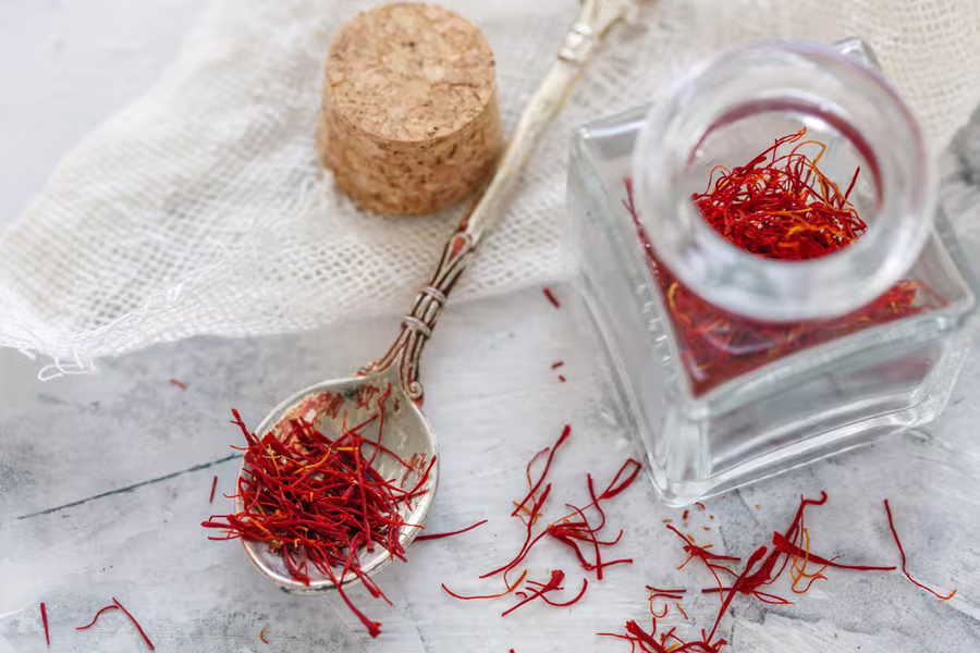 A silver spoon with red saffron threads next to a glass jar filled with saffron and a cork stopper, all on a white textured surface.